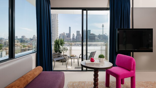 Habitación de hotel moderna con un balcón amplio, un sofá, una silla rosa, una mesita con un libro y un televisor.