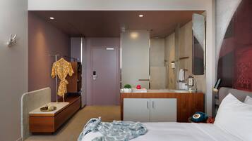 Hotelkamer met een bed, nachtkastje met lamp, bureau met stoel, garderobe en een badkamer met douche en spiegel.