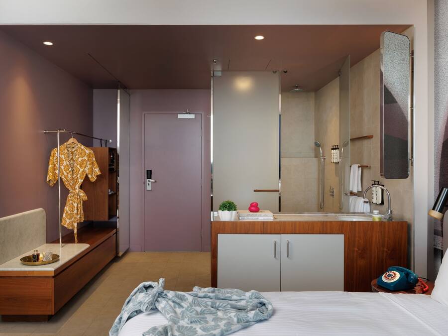 Hotelkamer met een bed, nachtkastje met lamp, bureau met stoel, garderobe en een badkamer met douche en spiegel.