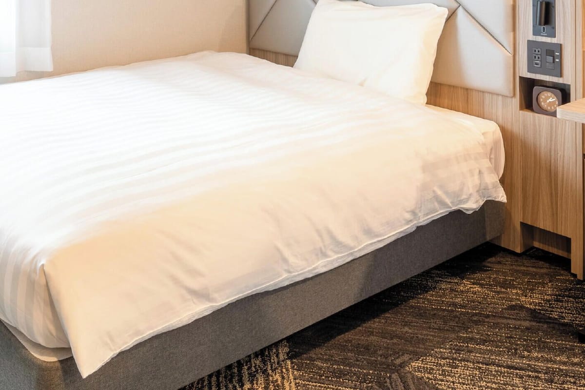 A neatly made bed with a grey headboard and a white pillow, a dark patterned carpet, and a light-colored wall.