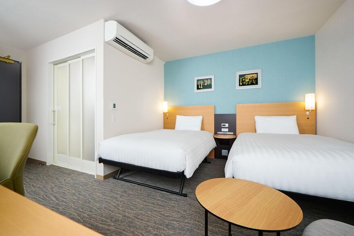 Hotellihuone, jossa on kaksi sänkyä, työpöytä, tuoli ja ilmastointilaite.