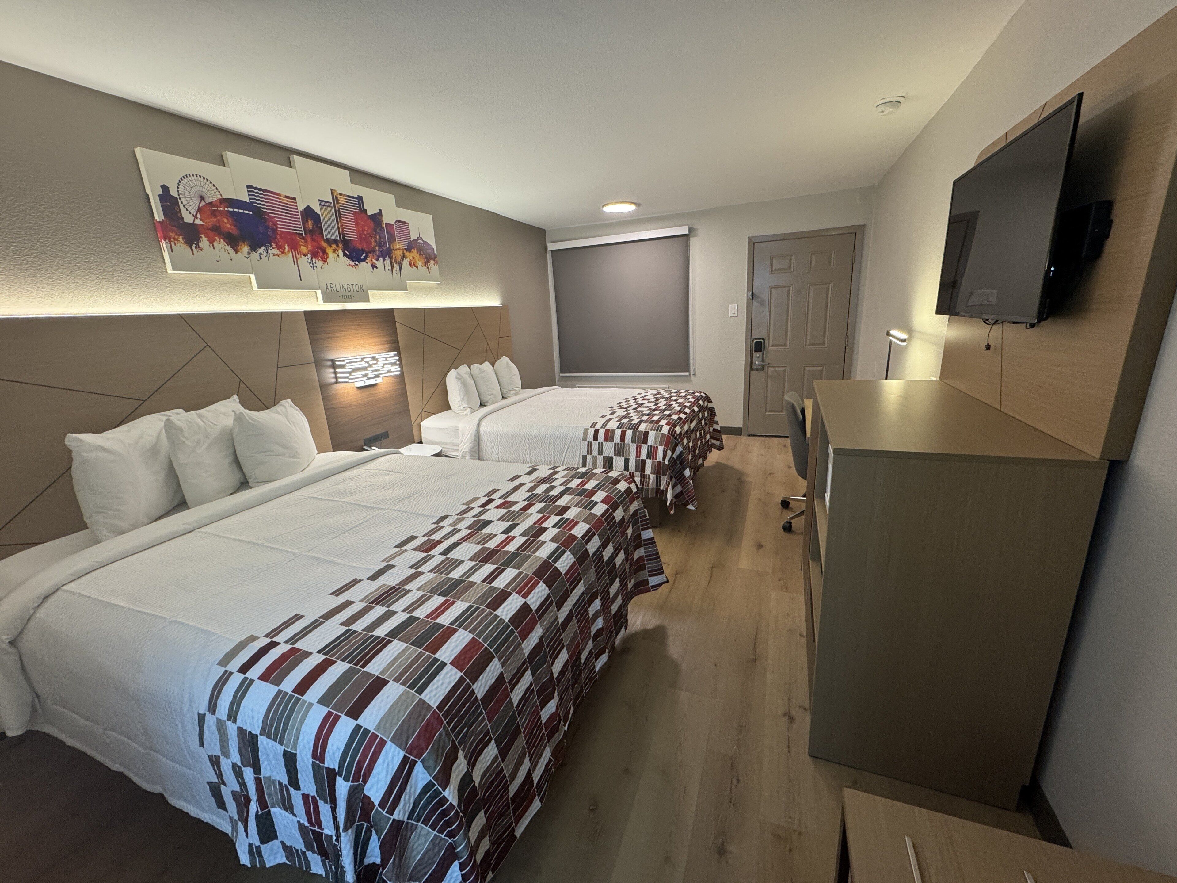 A hotel room with two beds, a TV mounted on the wall, a desk, and a painting of a city skyline.