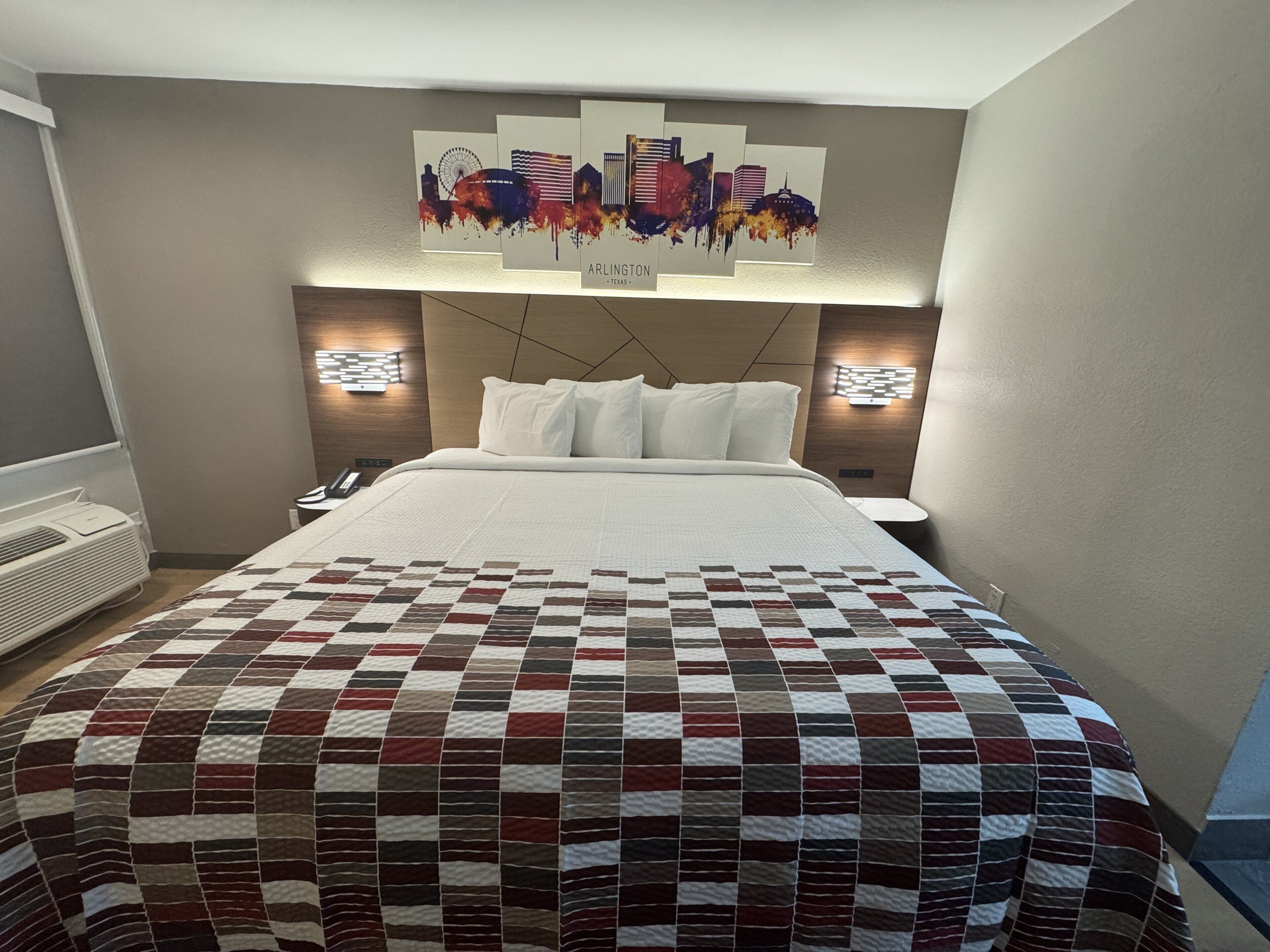 Une chambre d’hôtel avec un grand lit, une couvre-lit matelassée, deux lampes de chevet, une œuvre d’art fixée au mur représentant une vue panoramique de la ville, et un thermostat mural.