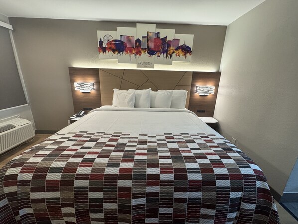 A hotel room with a large bed, a quilted bedspread, two bedside lamps, a wall-mounted artwork of a city skyline, and a thermostat on the wall.