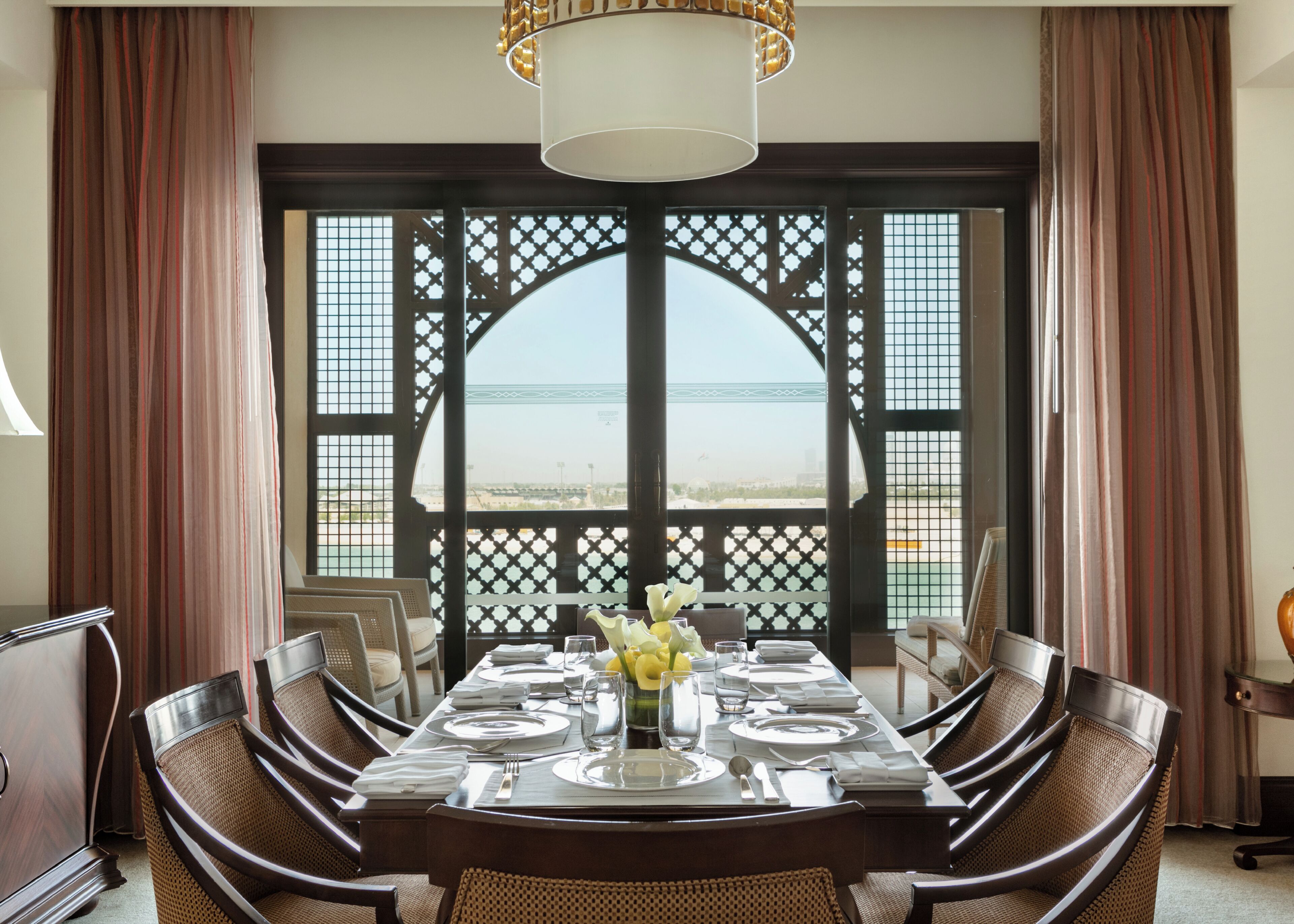 Un coin repas avec une table dressée pour six personnes, des chaises, un vase décoratif et une vue à travers une fenêtre à treillis.