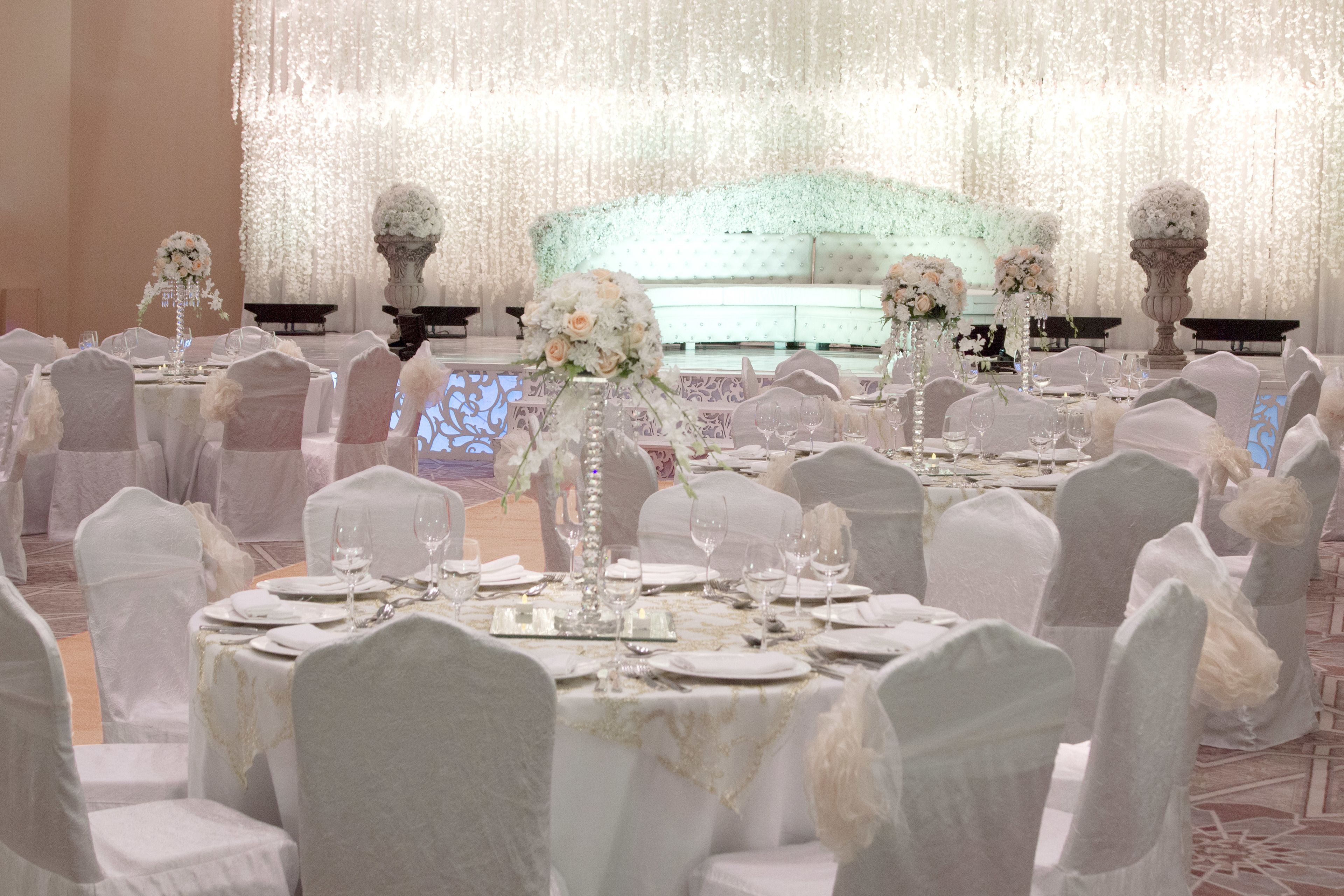 Salão de festas com mesas redondas preparadas para um evento formal, decorado com toalhas brancas e arranjos florais.
