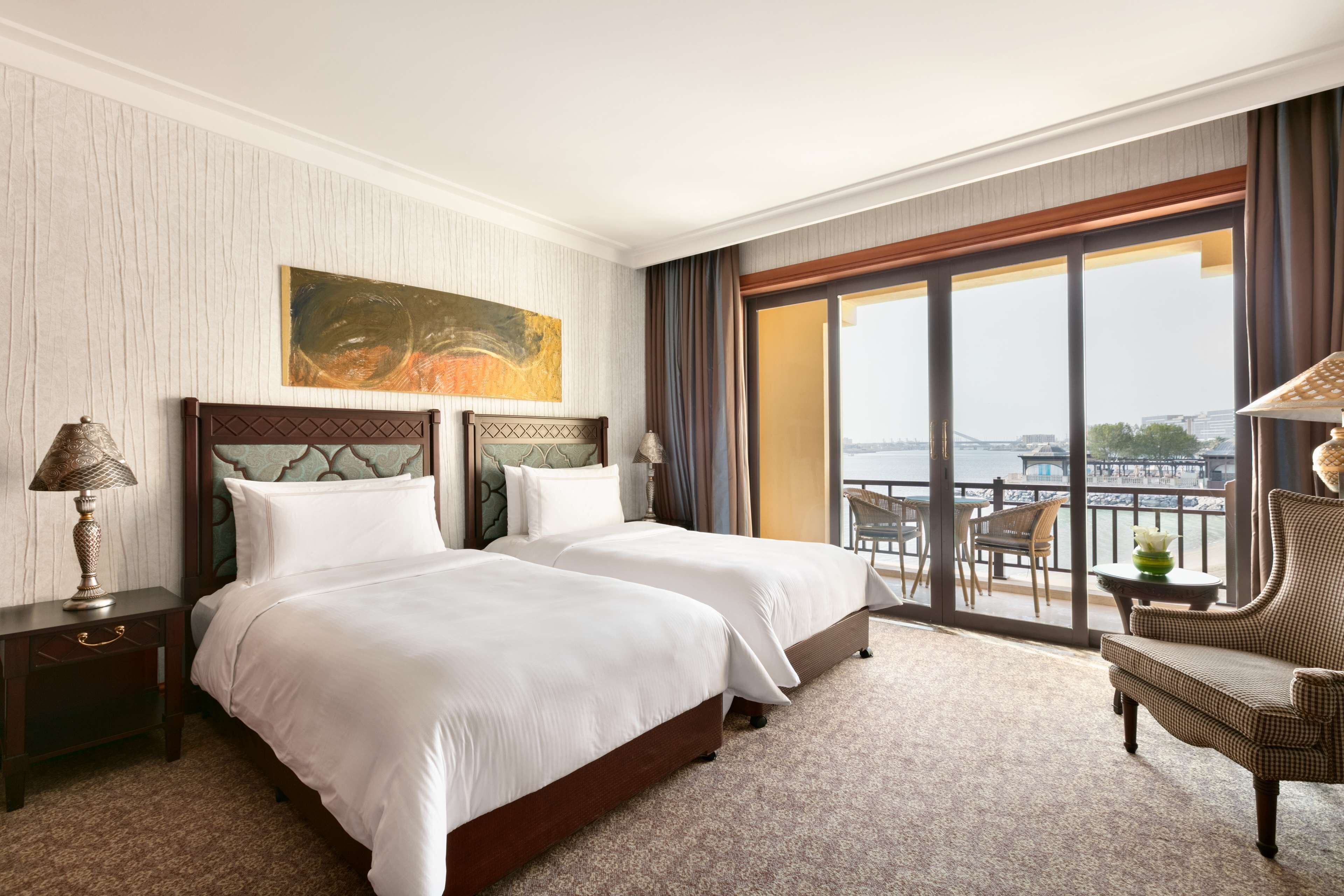 Une chambre d’hôtel avec deux lits, un balcon donnant sur un beau paysage, une chaise et un tableau au mur.