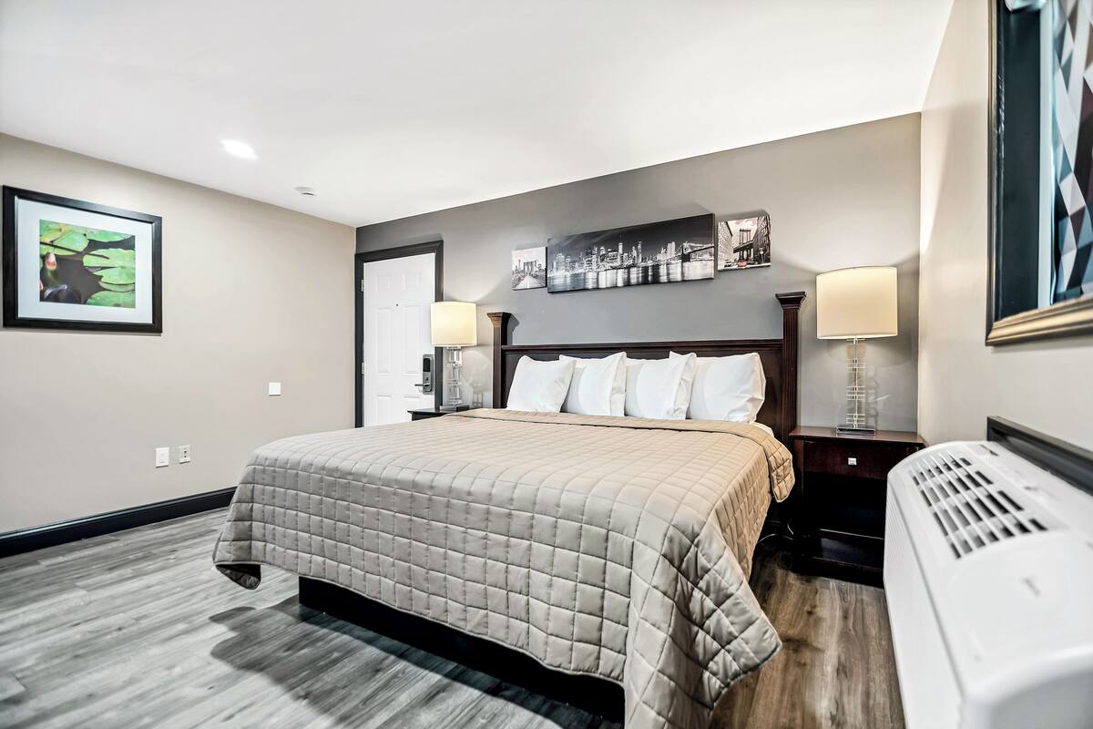 A hotel room with a bed, a nightstand, a lamp, a heater, and framed artwork on the walls.