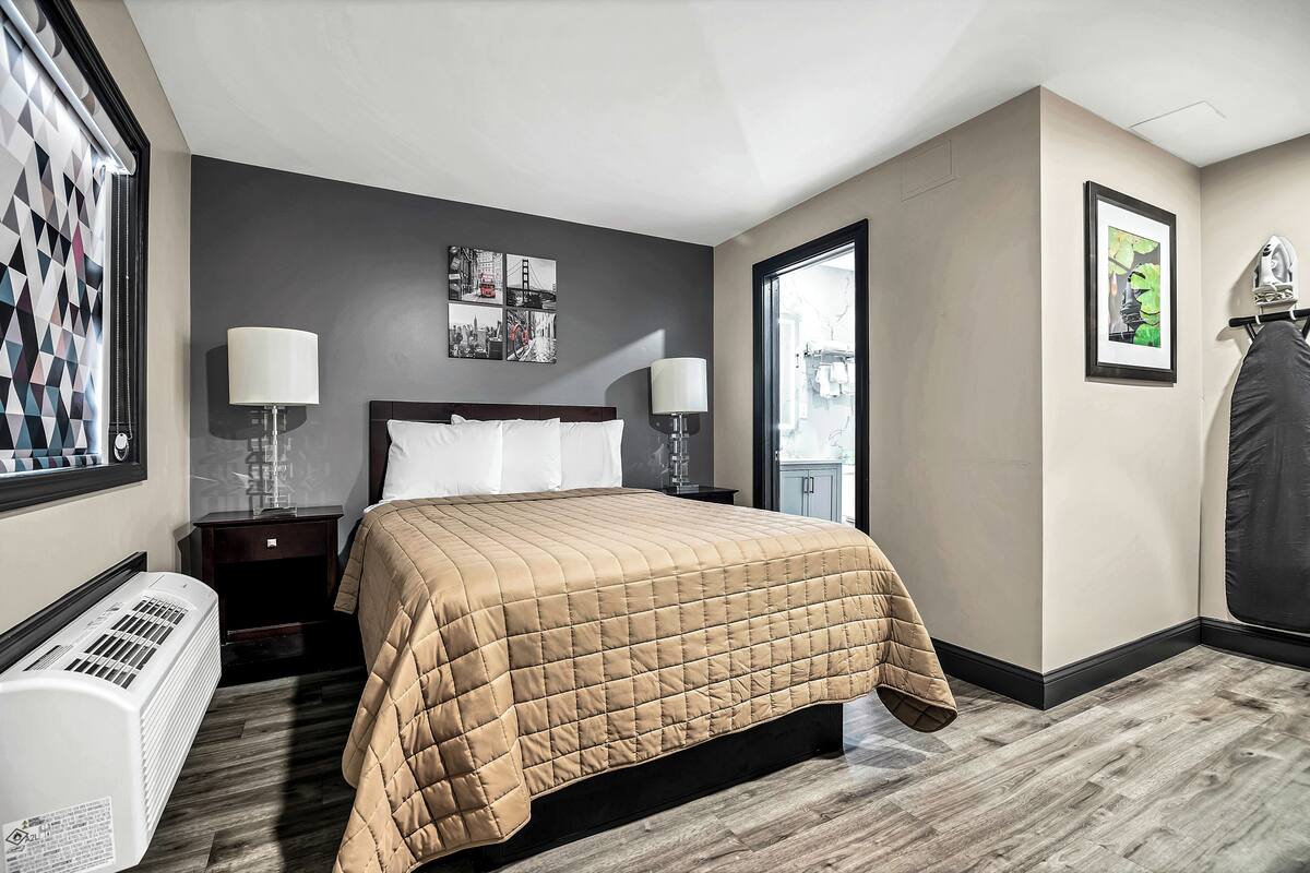 A hotel room with a bed, two bedside tables, a heater, a window with blinds, and a framed picture on the wall.