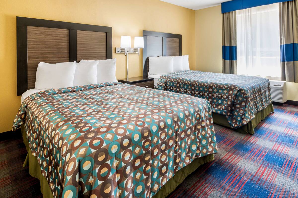 A hotel room with two beds, a large window with curtains, a nightstand, and a lamp.