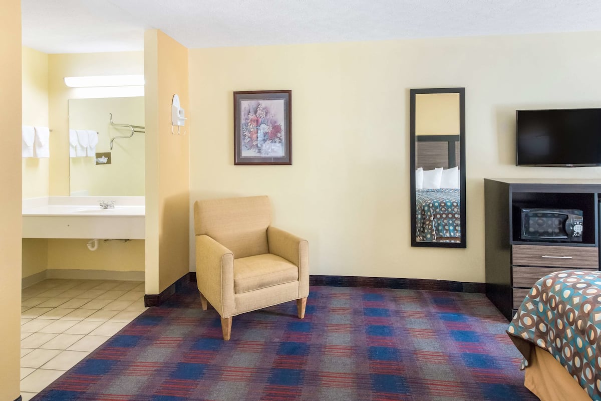 A hotel room with a bed, a chair, a television, a bathroom with a sink and mirror, and a framed picture on the wall.