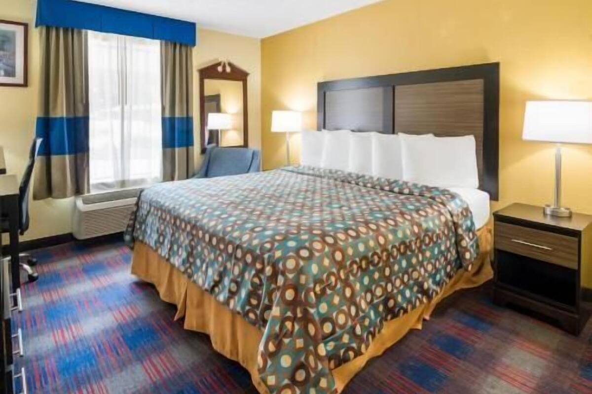 A hotel room with a bed, a desk, a chair, a lamp, and a window with curtains.