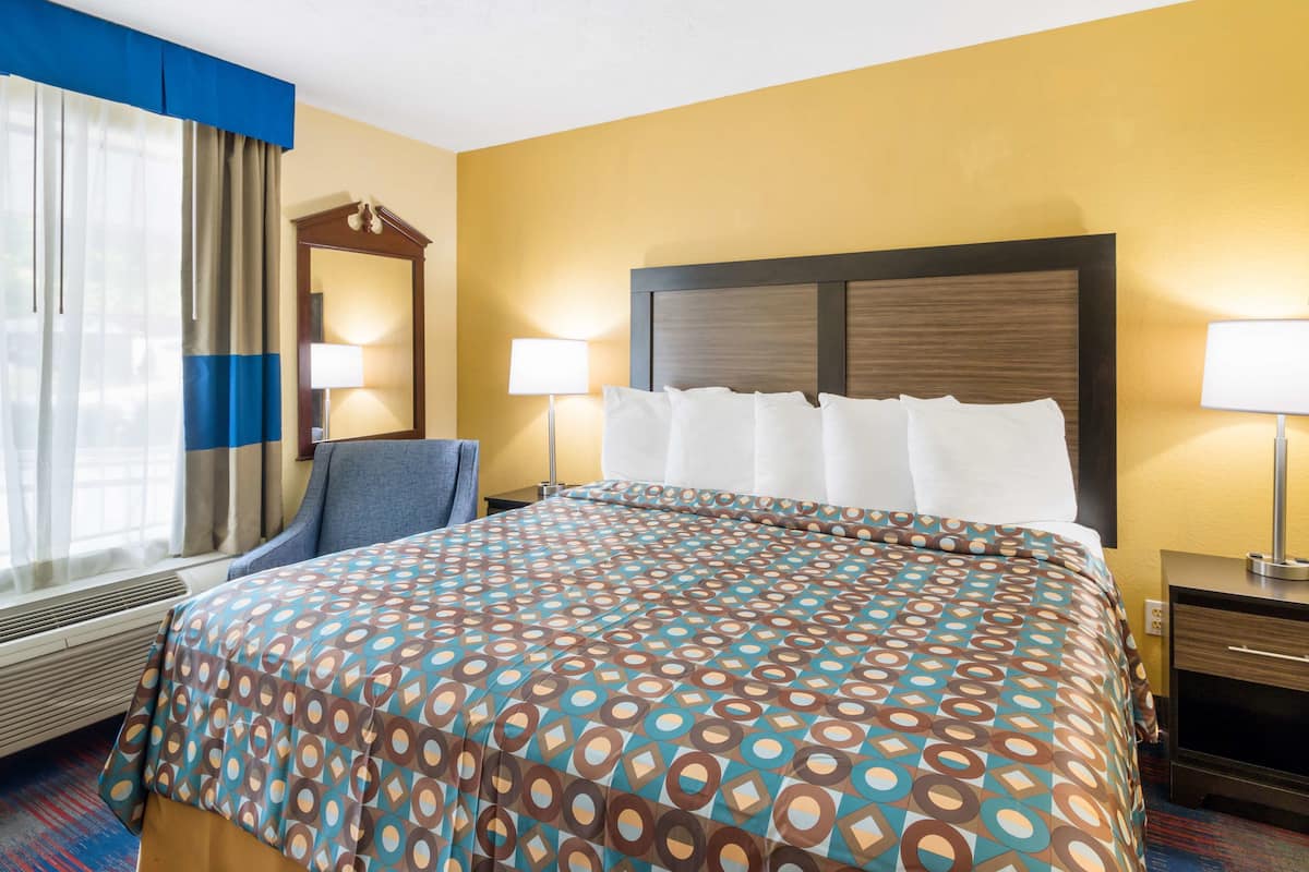 A hotel room with a bed, two bedside tables, a chair, a window with curtains, and a wall-mounted mirror.