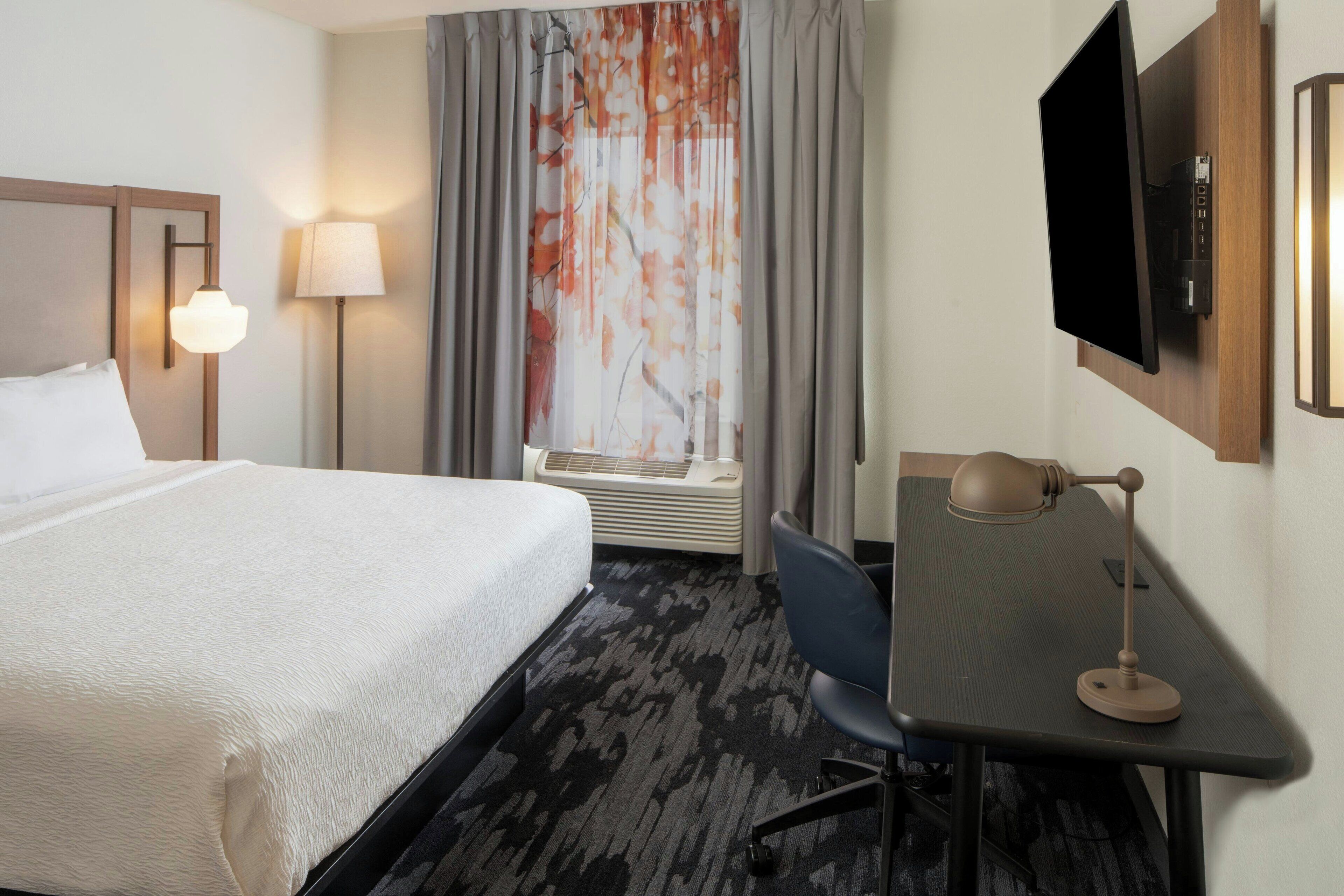 A hotel room with a bed, desk, chair, TV, lamp, and window with curtains.