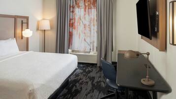 A hotel room with a bed, desk, chair, TV, lamp, and window with curtains.