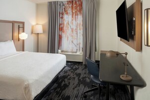 Habitación de hotel con cama, escritorio, silla, televisor, lámpara y ventana con cortinas.