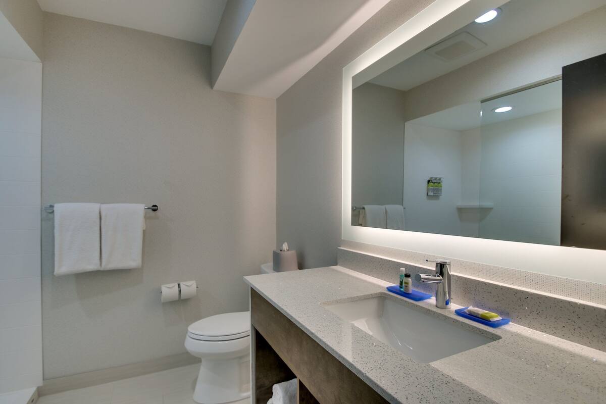 A modern bathroom with a large mirror, a white toilet, and a countertop with a sink and soap dispenser.