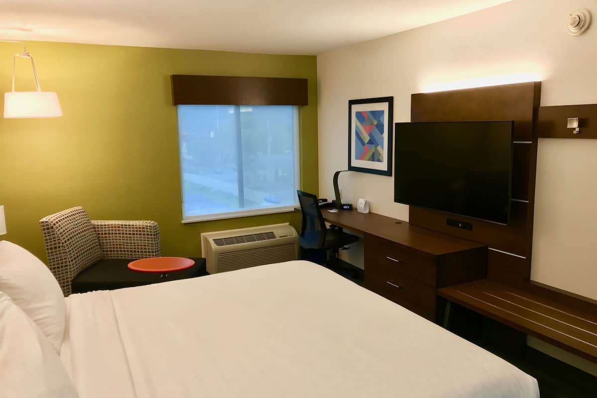 A hotel room with a bed, a desk with a computer, a chair, a TV, and a window with blinds.