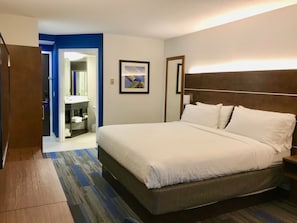 Una habitación de hotel con una cama grande, un baño con lavamanos y espejo, y una puerta que da a otra habitación.