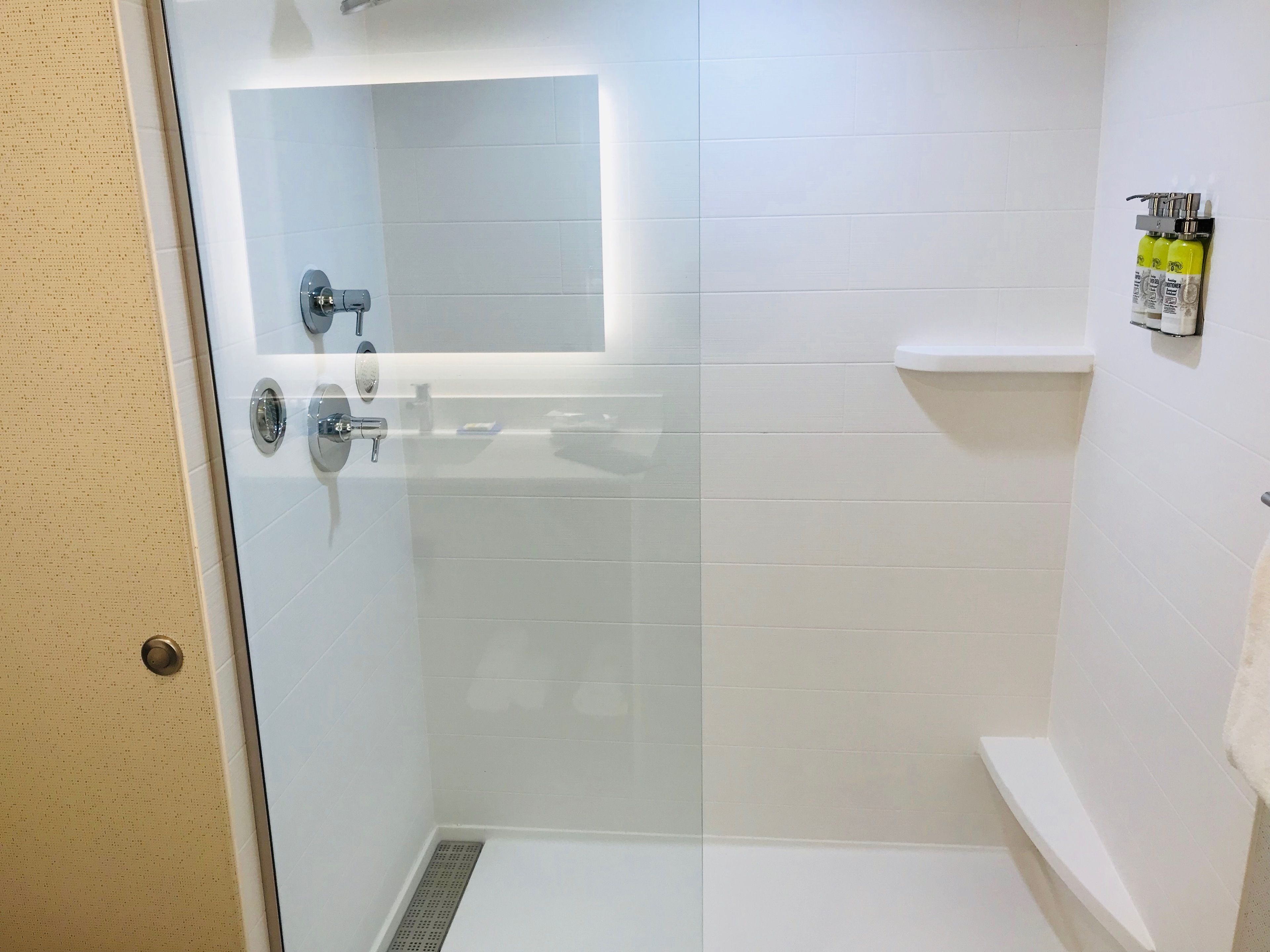 Une douche fermée par des parois de verre, avec des murs recouverts de carreaux blancs, un lavabo blanc et un porte-serviettes sur lequel sont posées des bouteilles.