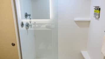 Une douche fermée par des parois de verre, avec des murs recouverts de carreaux blancs, un lavabo blanc et un porte-serviettes sur lequel sont posées des bouteilles.