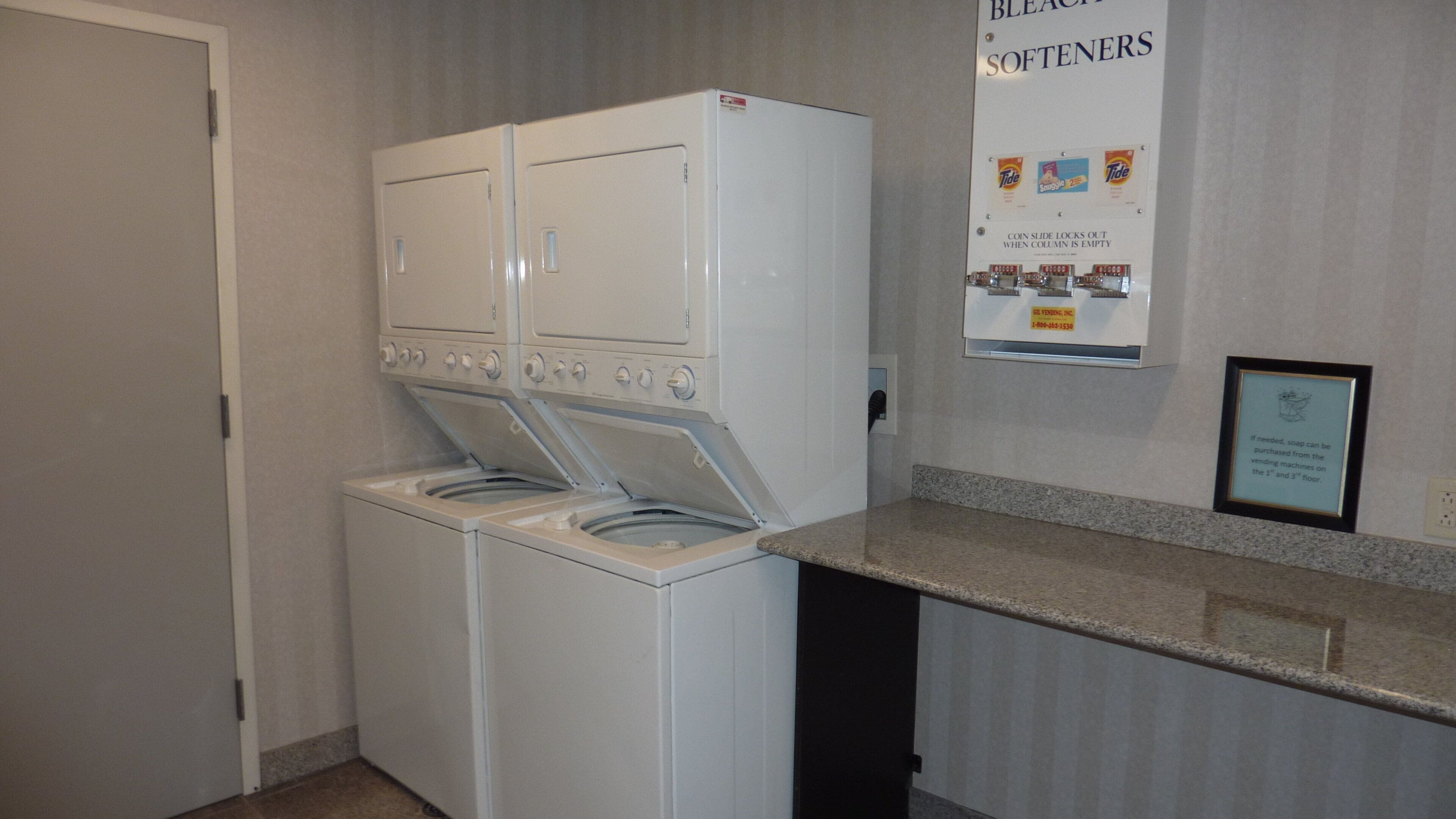 A laundry area with a front-loading washer and dryer, a coin-operated vending machine, and a sign indicating the location of softeners.