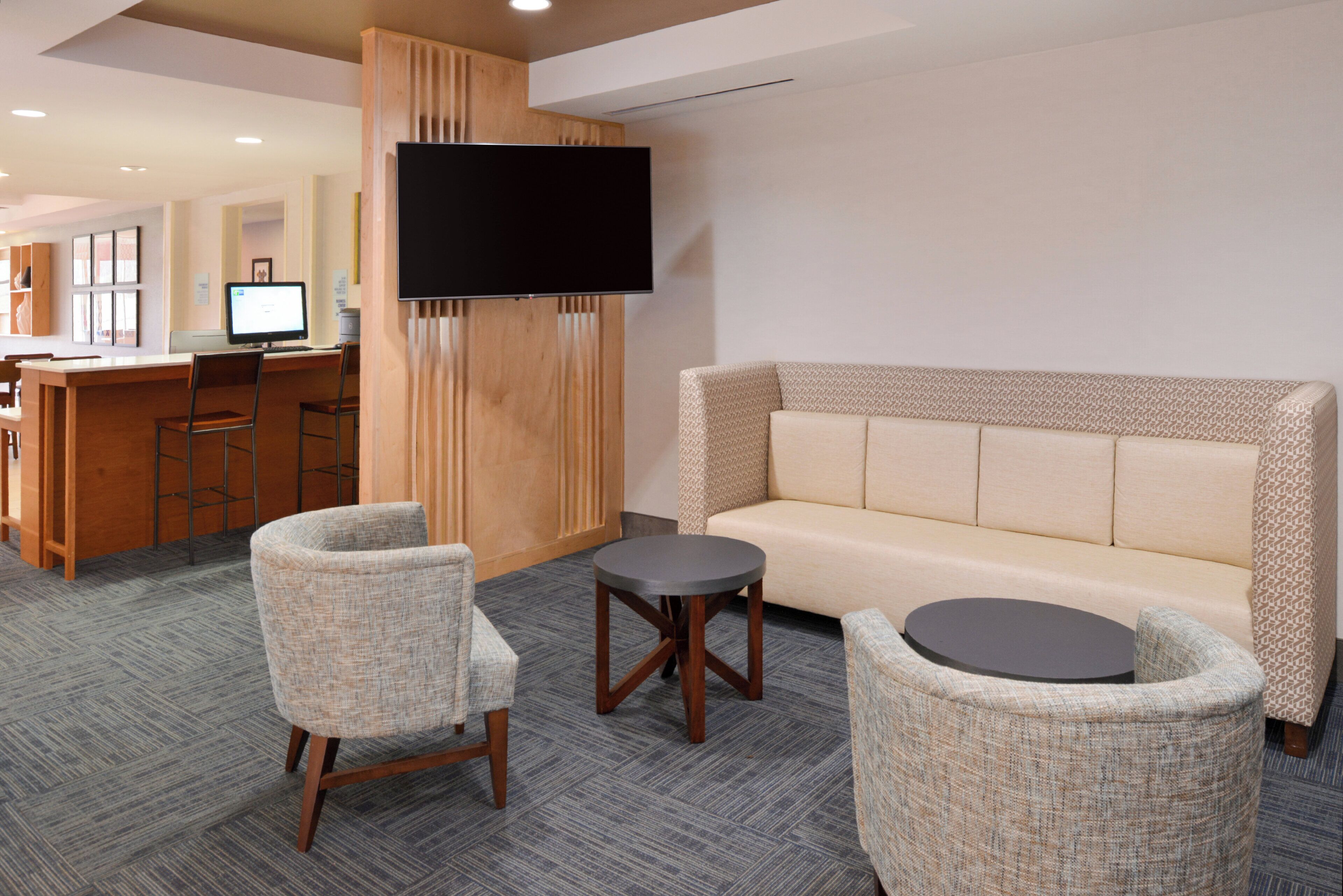 A hotel lobby with a sofa, two armchairs, a small table, a TV mounted on a wooden panel, and a desk with a computer.