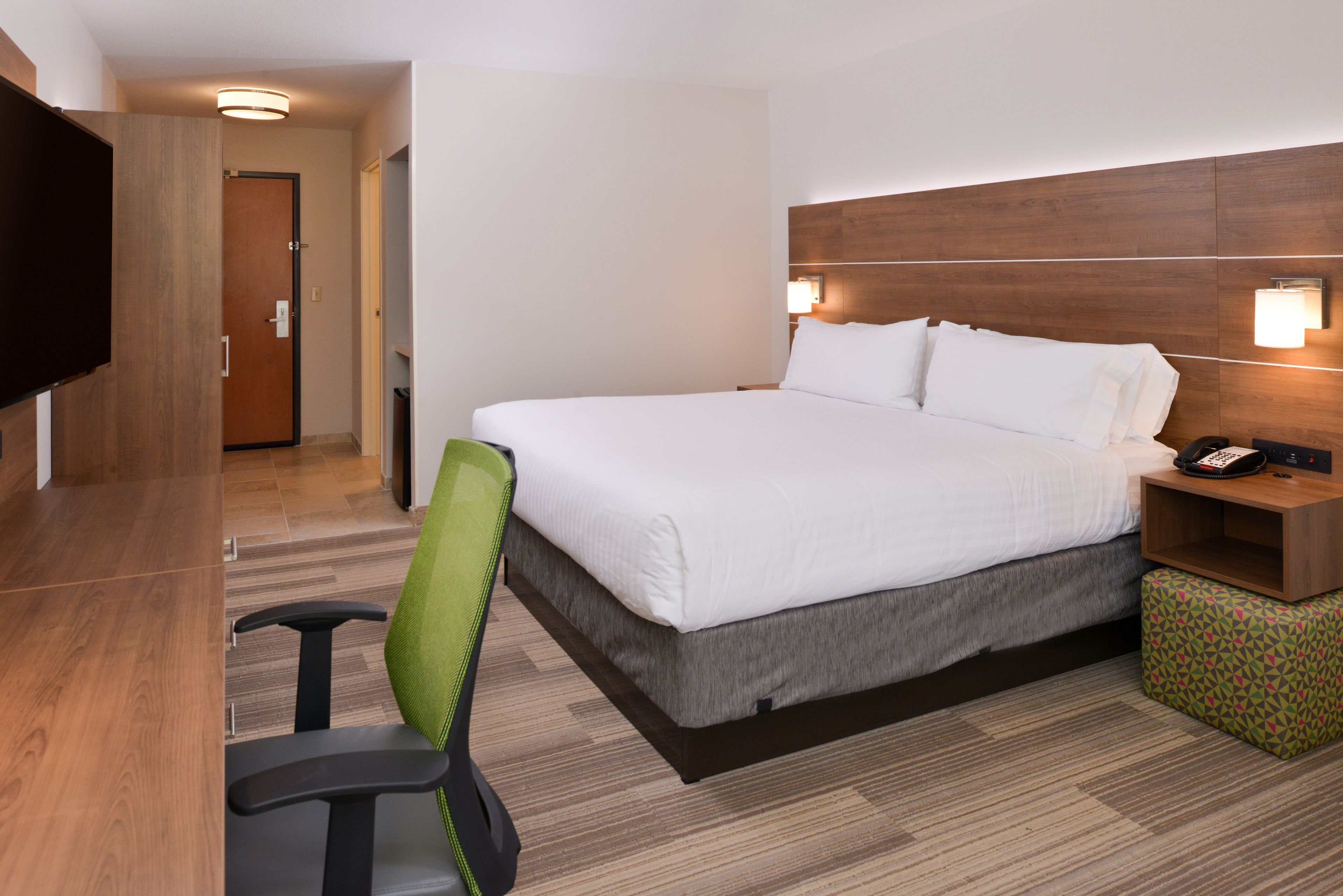 A hotel room with a bed, a desk, a chair, a TV, and a bedside table with a lamp.
