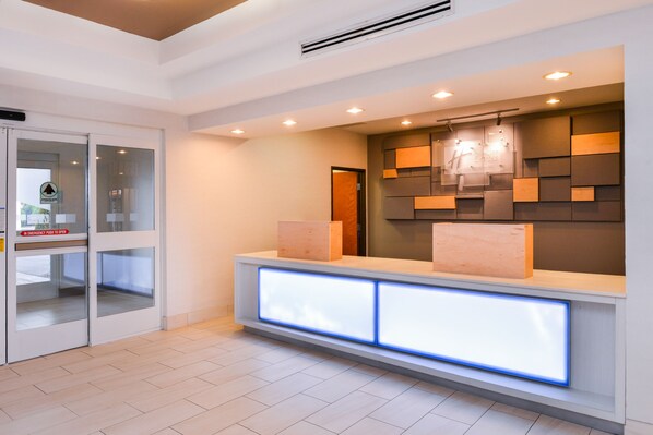 A reception area with a modern design, featuring a glass door entrance, a reception desk with illuminated panels, and a wall-mounted light fixture. - Holiday Inn Express & Suites Parkersburg-Mineral Wells by IHG (Mineralwells)