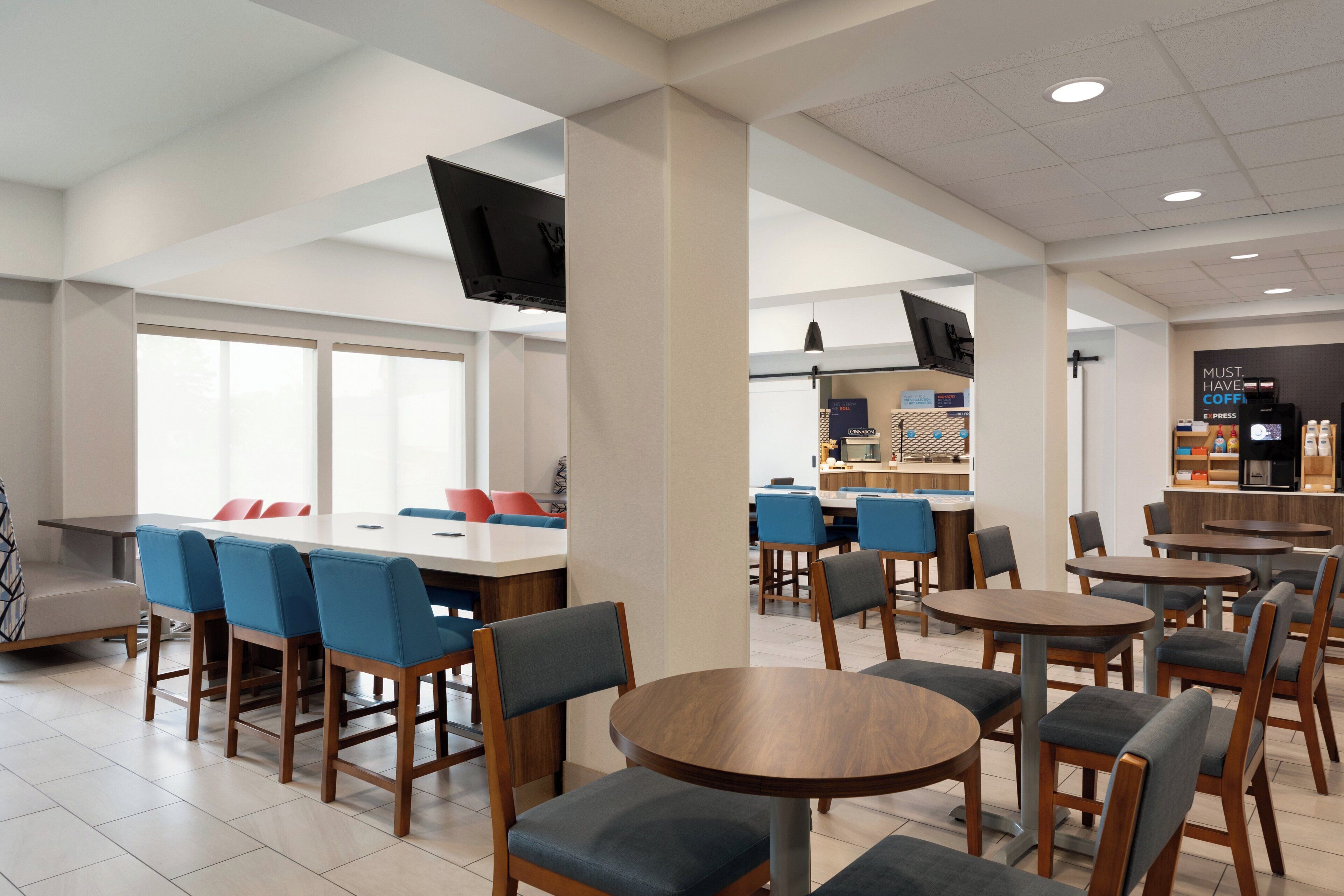 A modern cafe with round tables and blue chairs, a flat-screen TV mounted on a pillar, and a coffee bar in the background.
