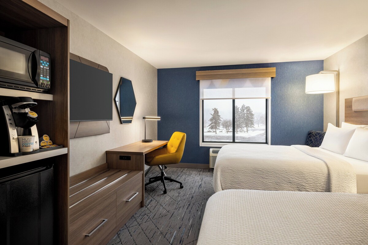 A hotel room with a bed, a desk with a chair, a microwave, a coffee maker, and a window with a view of trees.