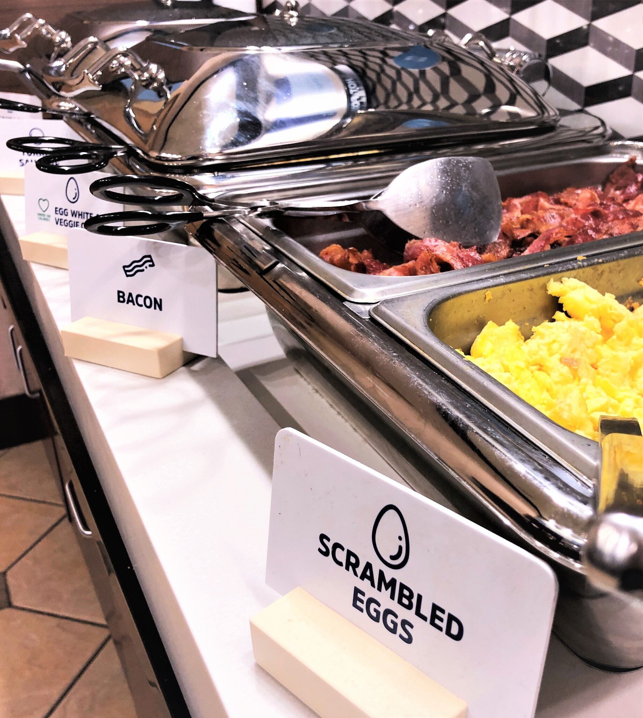 A buffet with labeled sections for scrambled eggs, bacon, and other dishes.