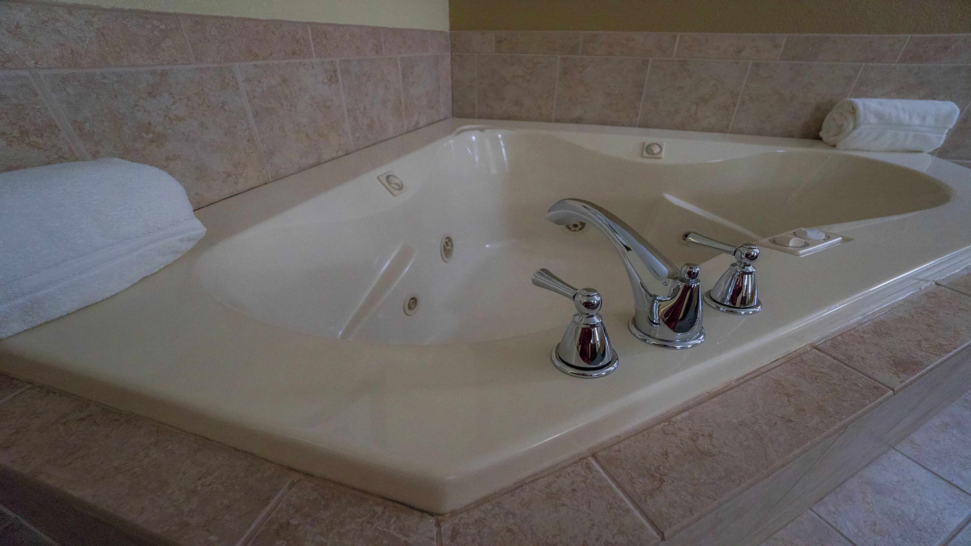A large bathtub with two faucets and a single control knob, surrounded by beige tiles.