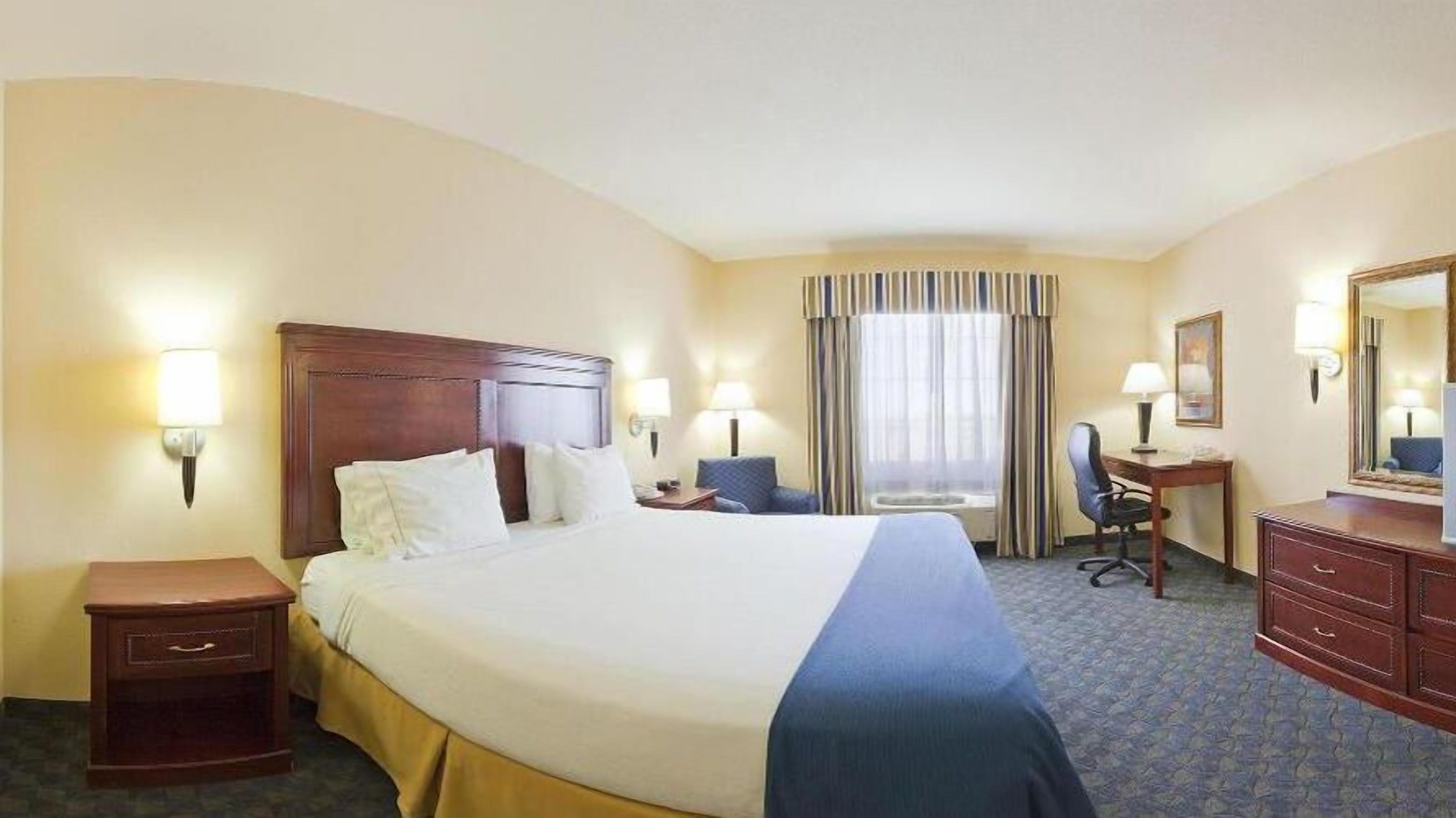 A hotel room with a large bed, bedside tables, a desk, a chair, a dresser, and a window with curtains.