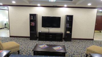 Habitación de hotel con un televisor de pantalla plana montado en la pared, flanqueado por dos estanterías. Hay una pequeña mesa con revistas frente a un sofá.