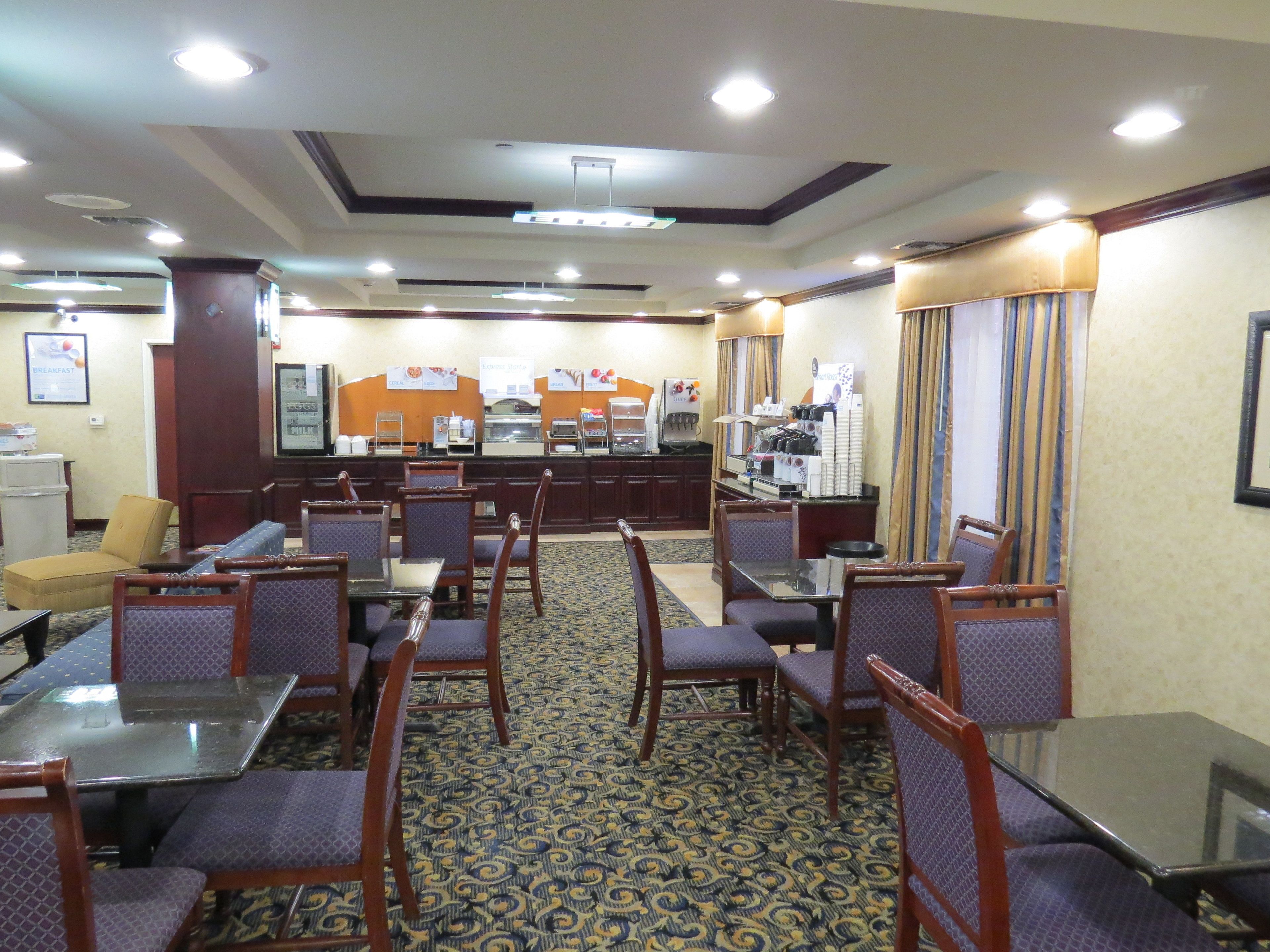 Un comedor de hotel con varias mesas y sillas, una barra con opciones de comida y bebidas, y una alfombra estampada.