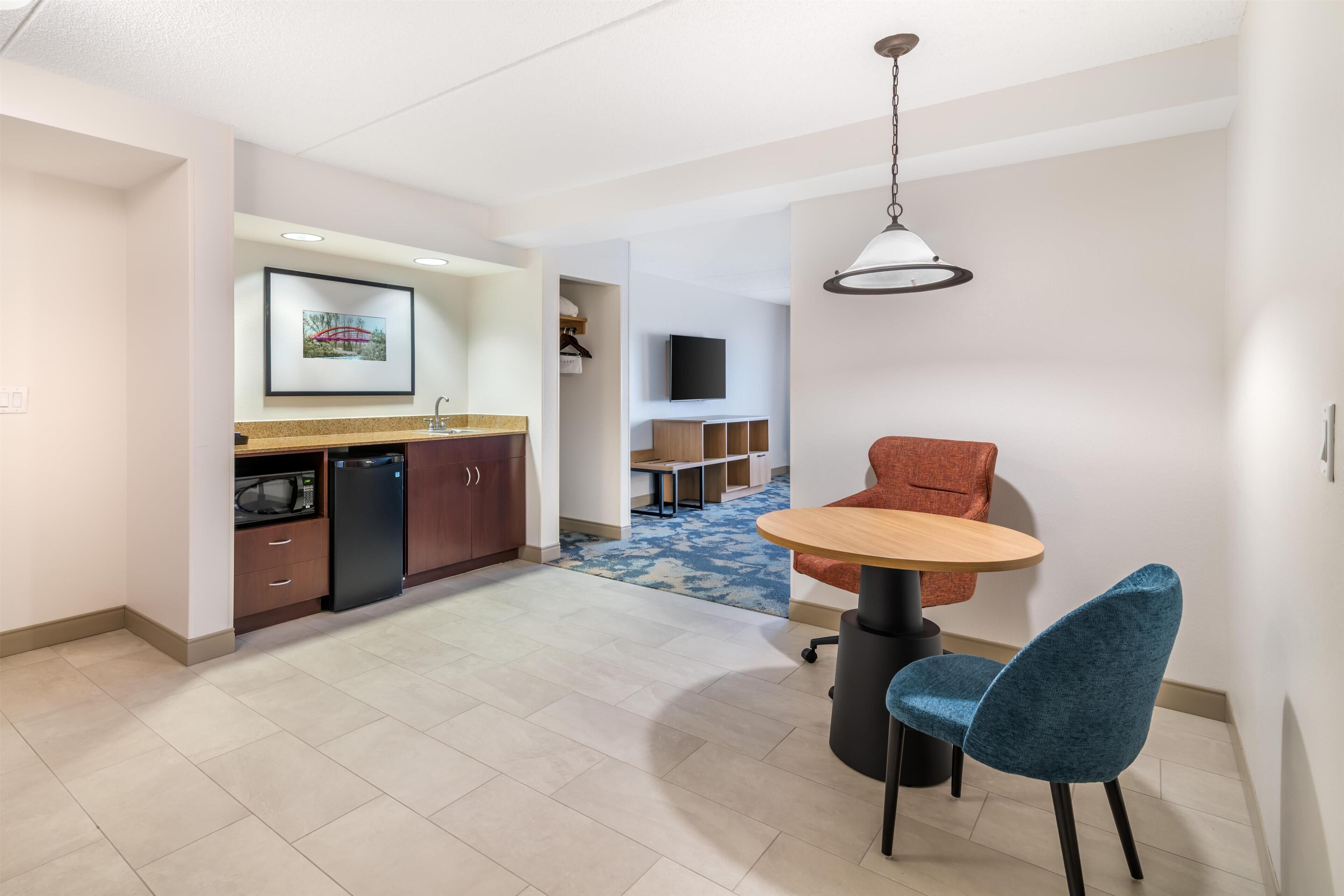 A modern kitchen with a microwave, refrigerator, and wooden cabinets. There is a dining area with a round table and two chairs, and a television mounted on the wall.