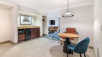 A modern kitchen with a microwave, refrigerator, and wooden cabinets. There is a dining area with a round table and two chairs, and a television mounted on the wall.