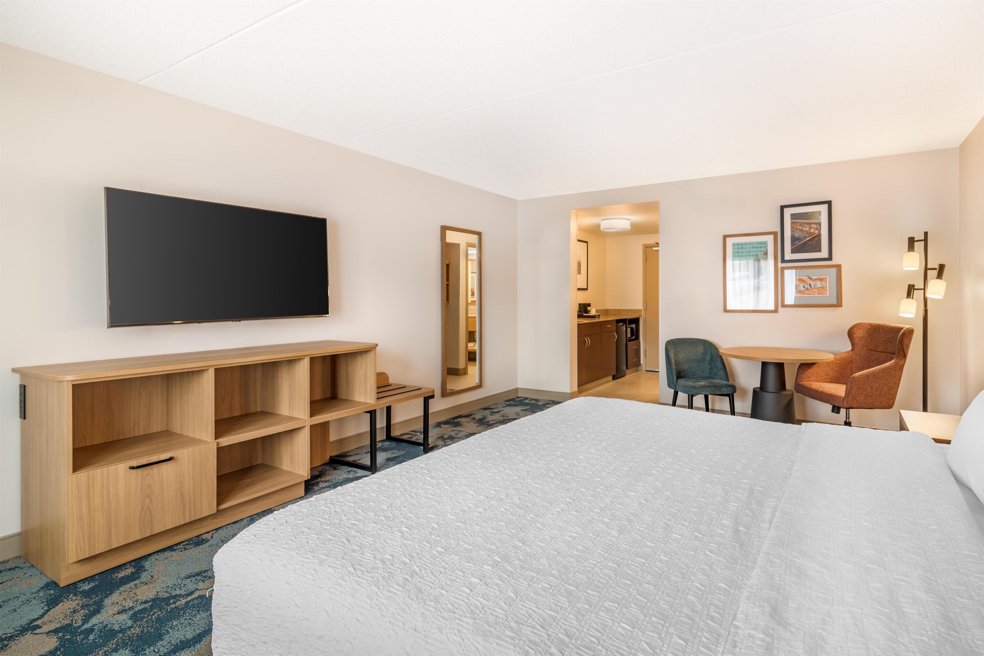 A hotel room with a large bed, a TV mounted on the wall, a wooden cabinet, a desk with a chair, and a mirror.