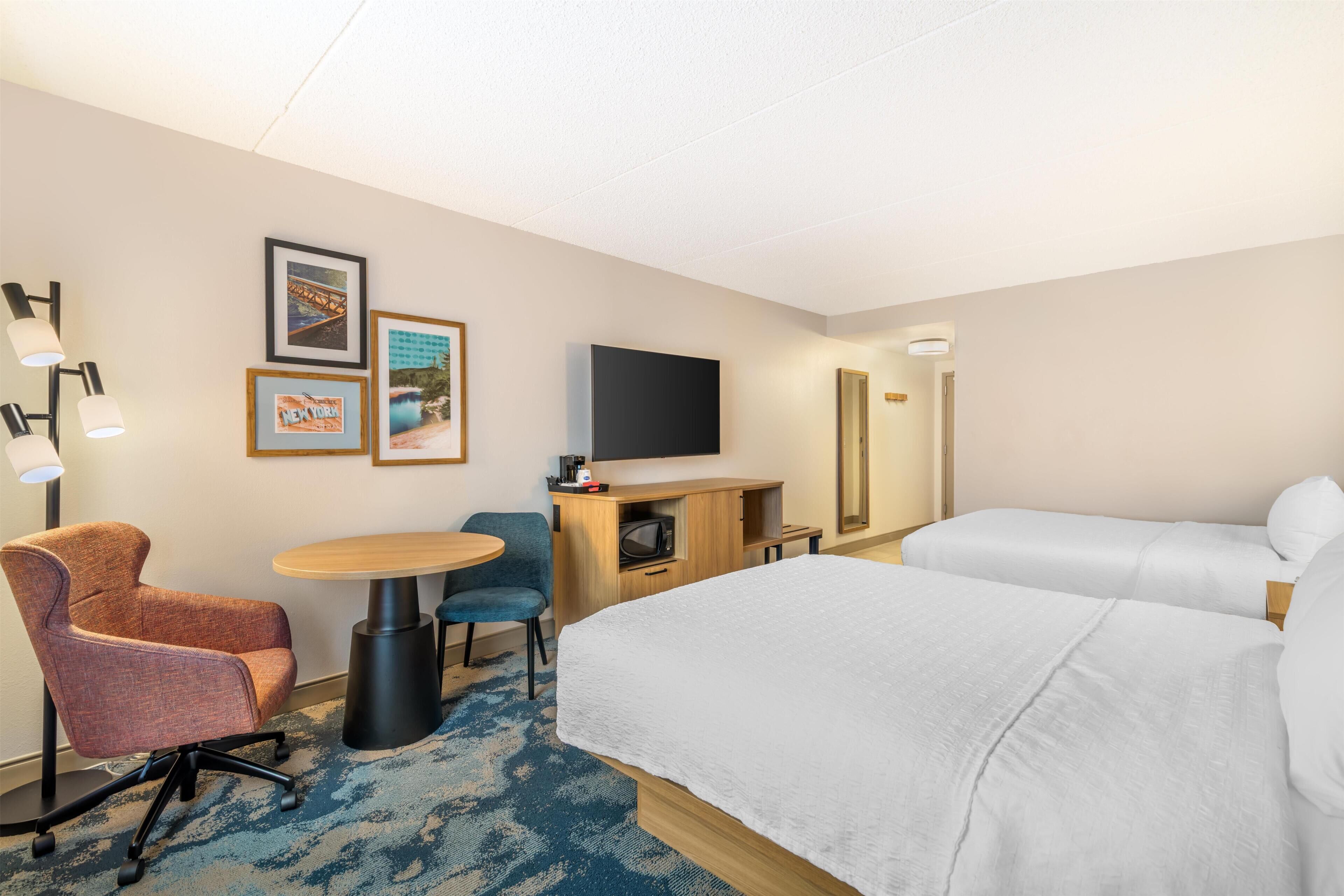 A hotel room with a bed, a desk with a computer, a chair, a TV mounted on the wall, and framed pictures on the wall.