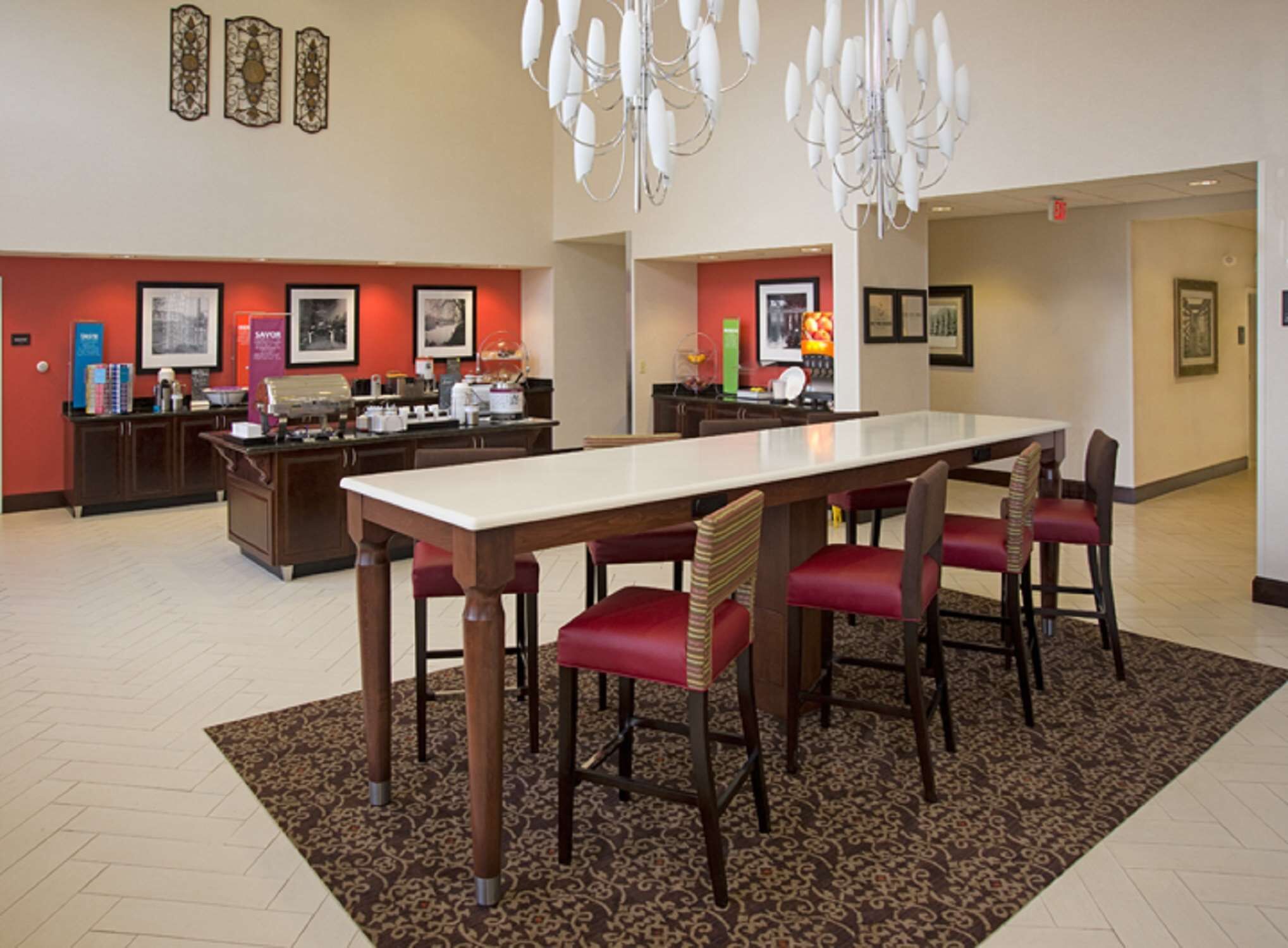 Un comedor de hotel con una larga mesa, sillas tapizadas en rojo, una alfombra estampada y un candelabro.