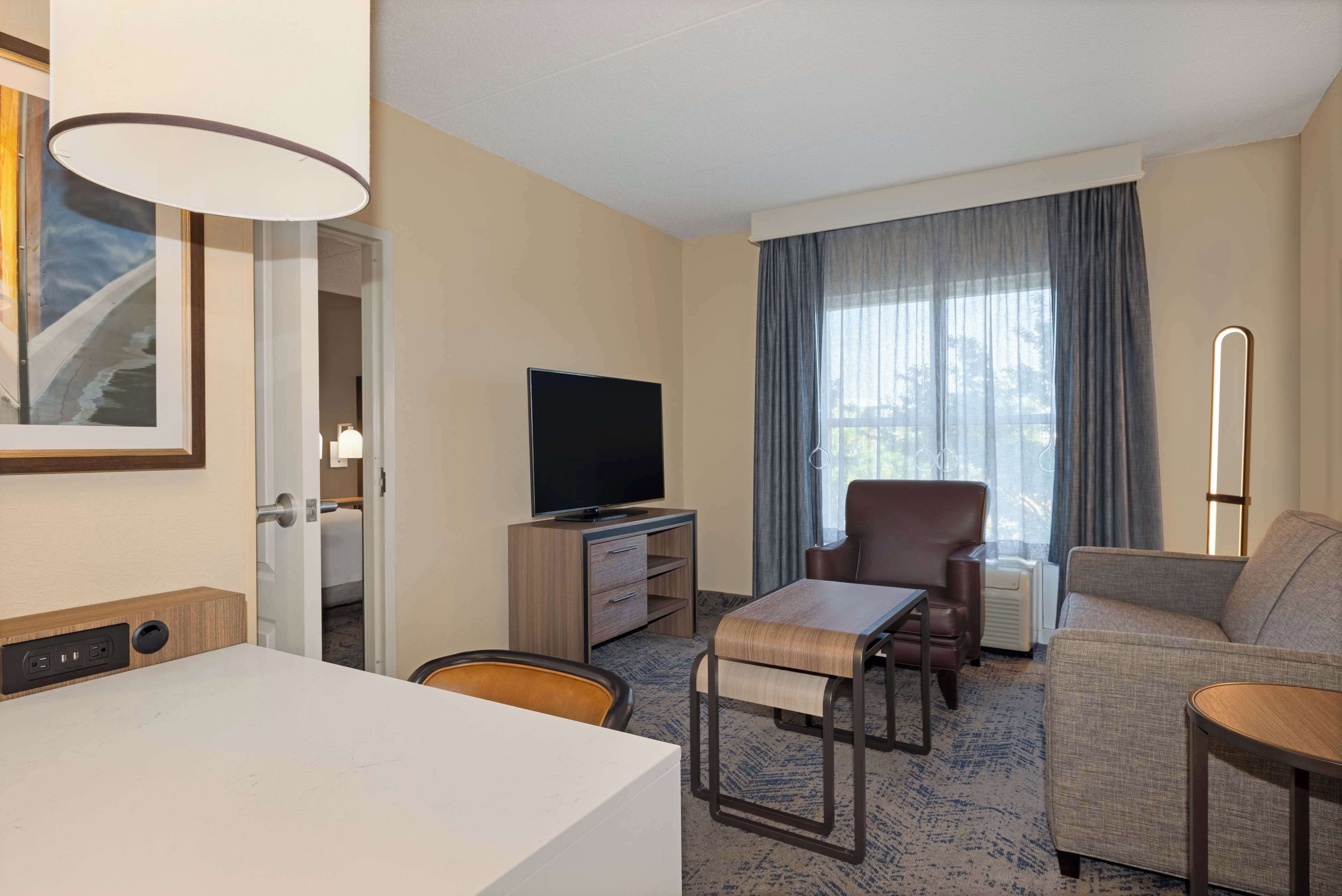 A hotel room with a flat-screen TV, a sofa, a chair, a small table, and a lamp.