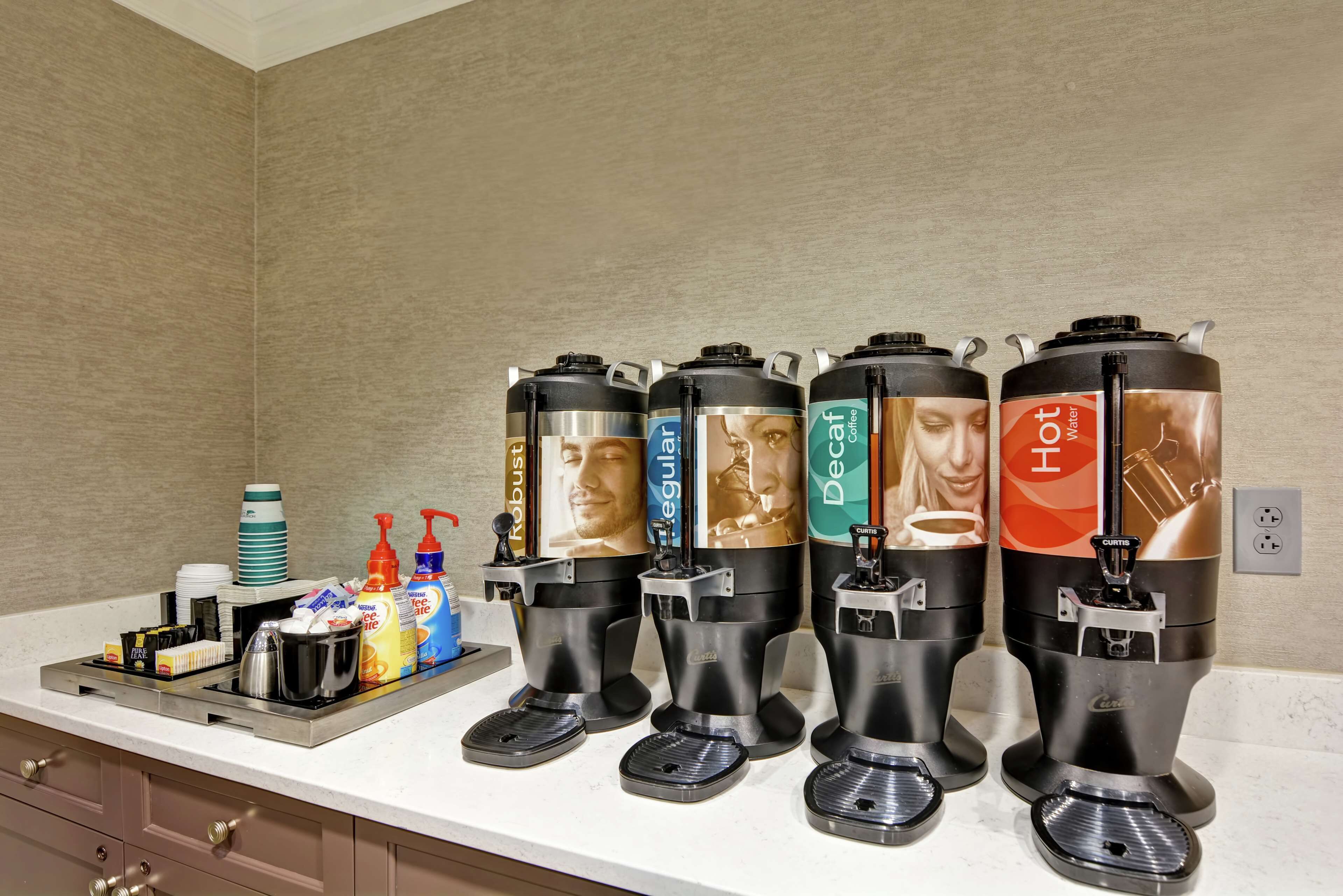 A coffee station with four dispensers labeled 'Robust', 'Regular', 'Decaf', and 'Hot Water'. There are also coffee cups, a coffee machine, and condiments on the counter.
