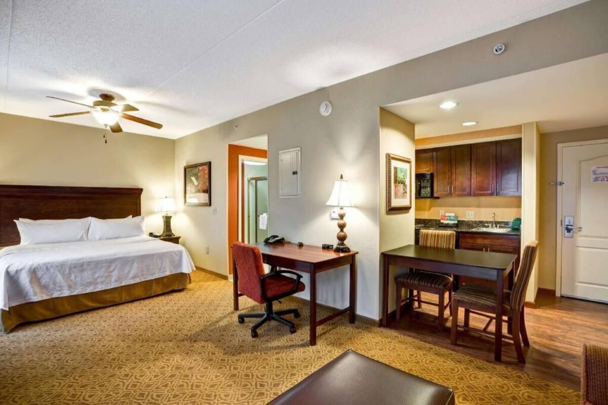 Une chambre d’hôtel équipée d’un lit, d’un bureau, d’une chaise, d’une kitchenette et d’un ventilateur de plafond.