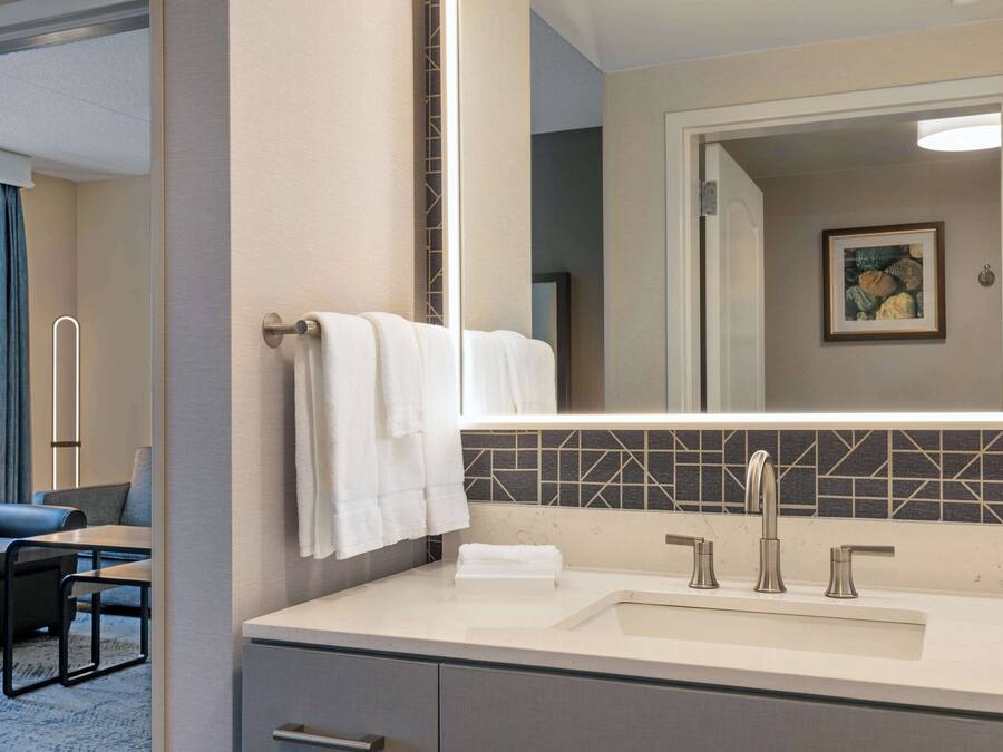 A hotel bathroom with a large mirror, a sink with two faucets, and a towel rack with two towels.