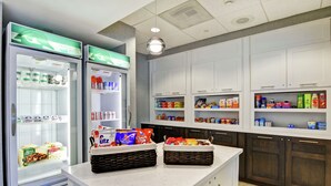 A kitchen area with two refrigerators stocked with food items, a countertop with baskets of snacks, and cabinets above holding various food products.