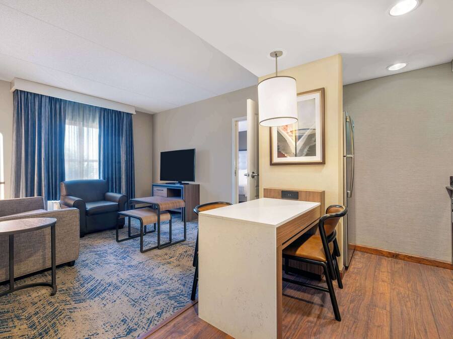 Habitación de hotel moderna con sala de estar, mesa de comedor, cocina y un amplio ventanal con cortinas.