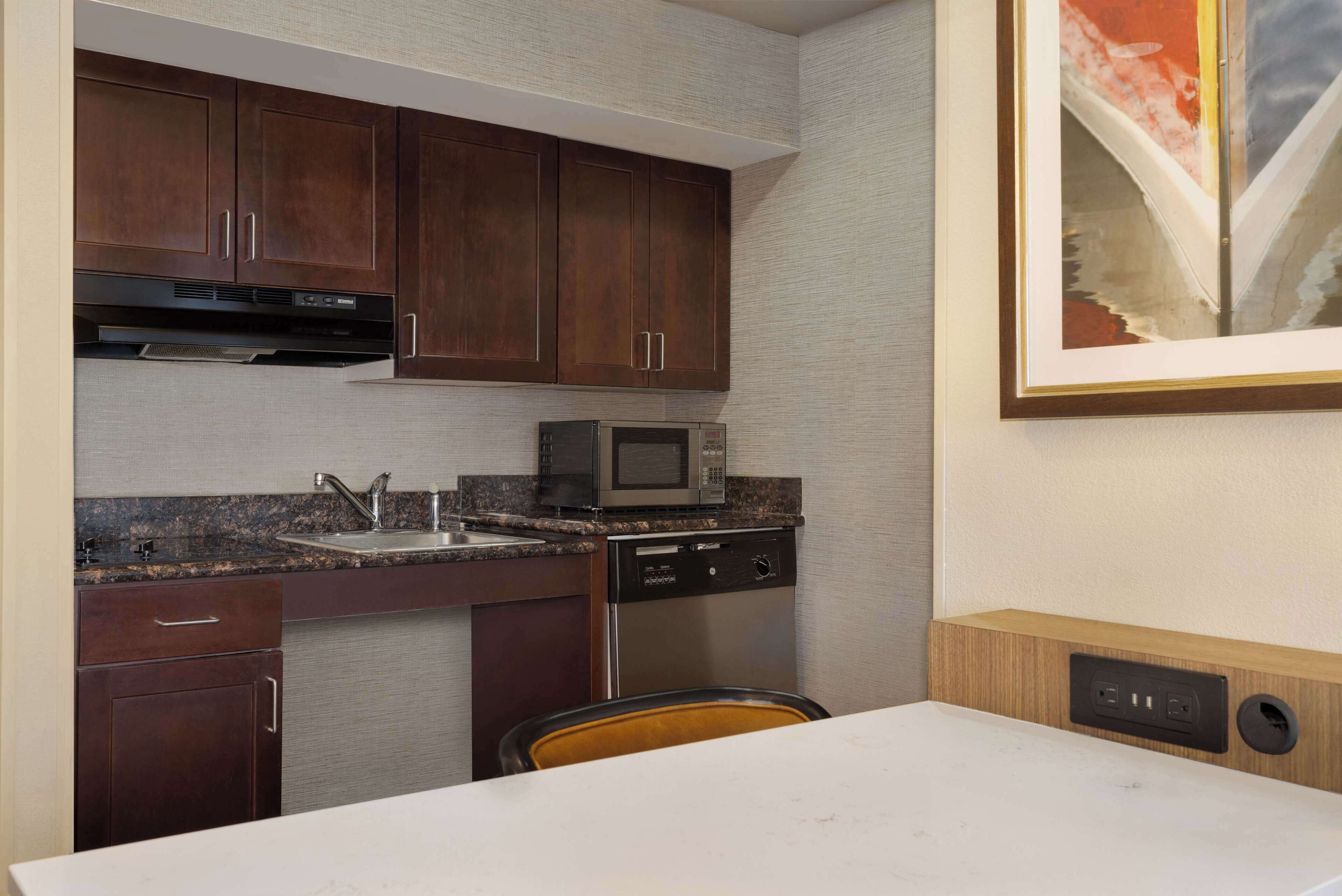 A compact kitchen with dark wooden cabinets, a stainless steel dishwasher, a microwave, and a granite countertop.