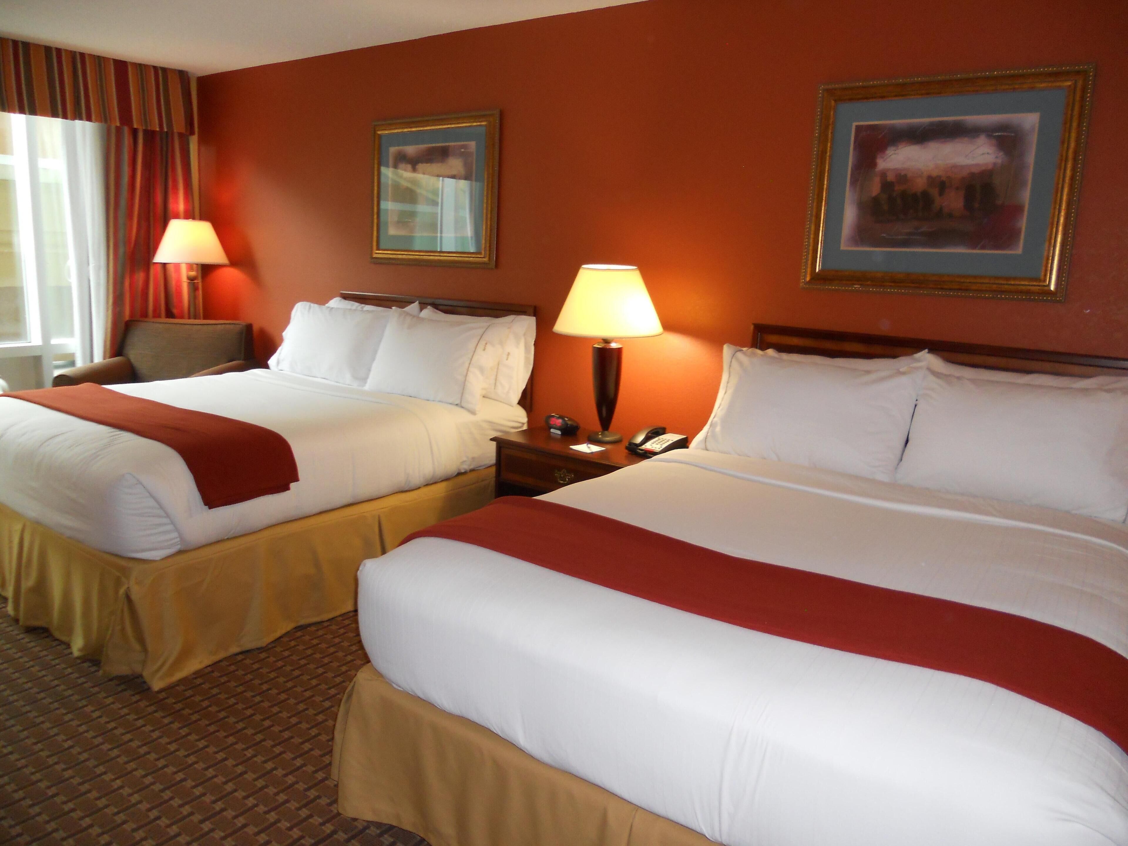 A hotel room with two beds, each with white linens and red throw blankets, a wooden headboard, a nightstand with a lamp, and a framed picture on the wall.