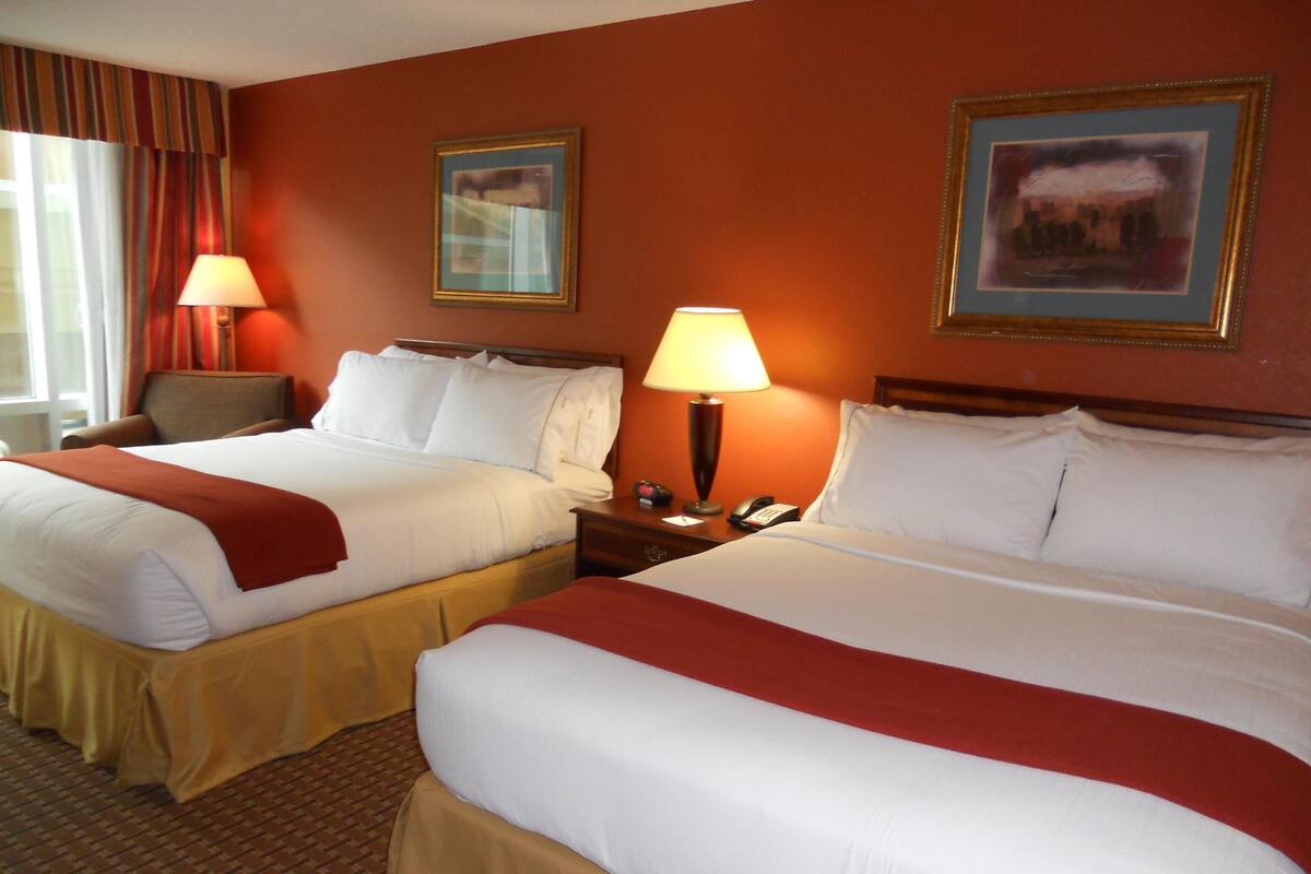 A hotel room with two beds, each with white linens and red throw blankets, a wooden headboard, a nightstand with a lamp, and a framed picture on the wall.